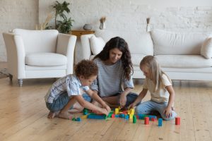Young mother her children, preschoolers son and daughter sit on warm floor in living room play together colored wooden blocks, spend leisure, carefree weekend at home. Kids development, games concept
