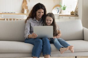 Positive happy young mother and sweet preschool kid girl using computer on home sofa, watching cartoon movie on laptop, hugging, enjoying domestic online internet communication, leisure